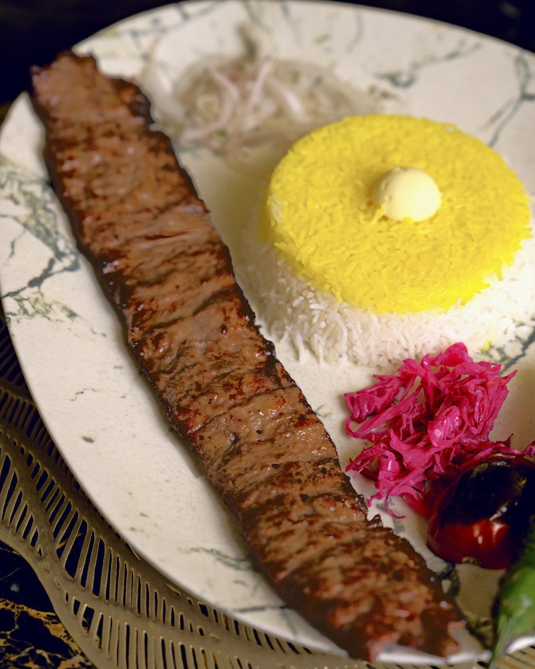 Barg Kebab beside saffron rice, red pickled onion and a grilled tomato on a marble-veined plate