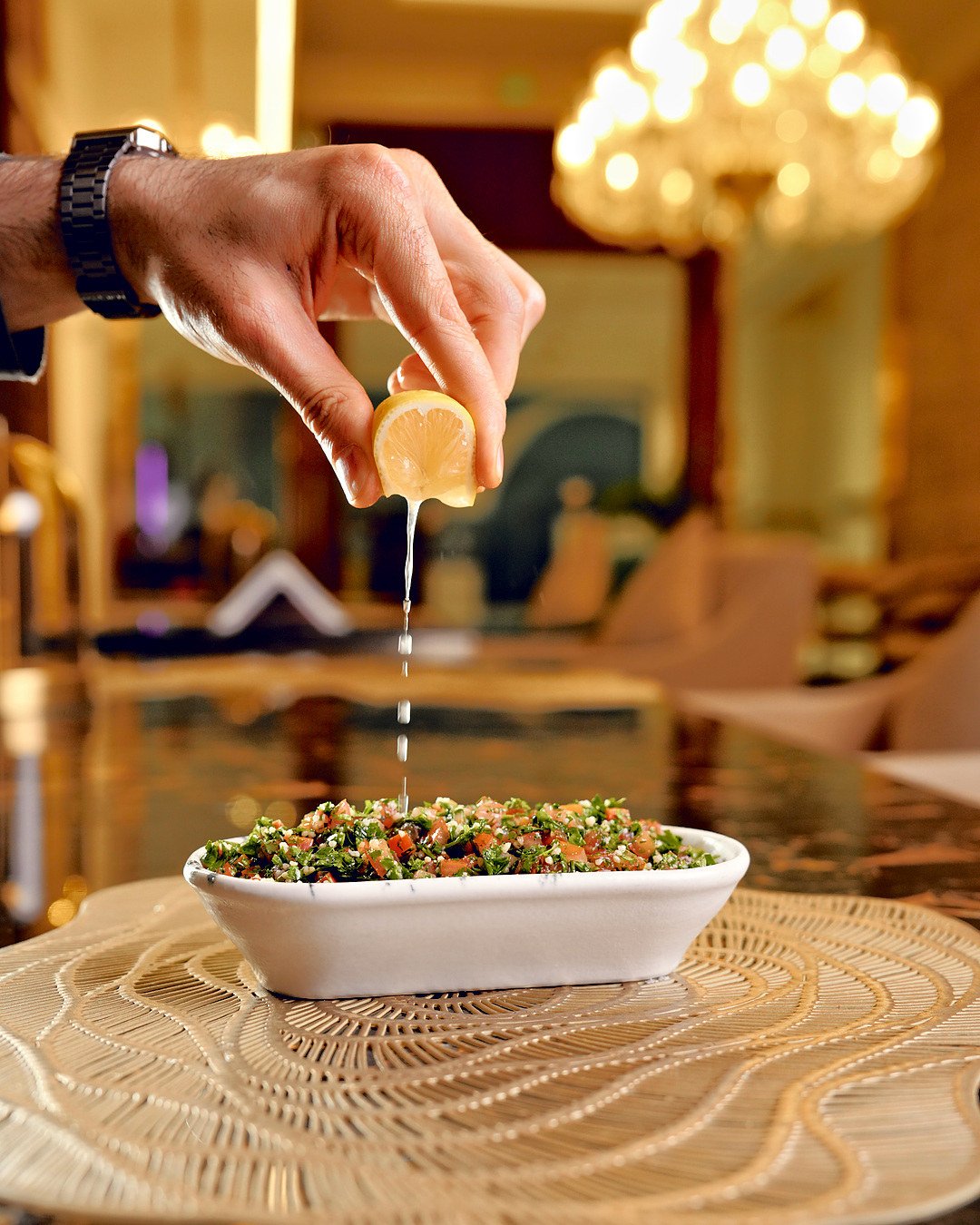 A lemon squeezed over Tabbouleh in a white dish, the chandeliers of the hall behind
